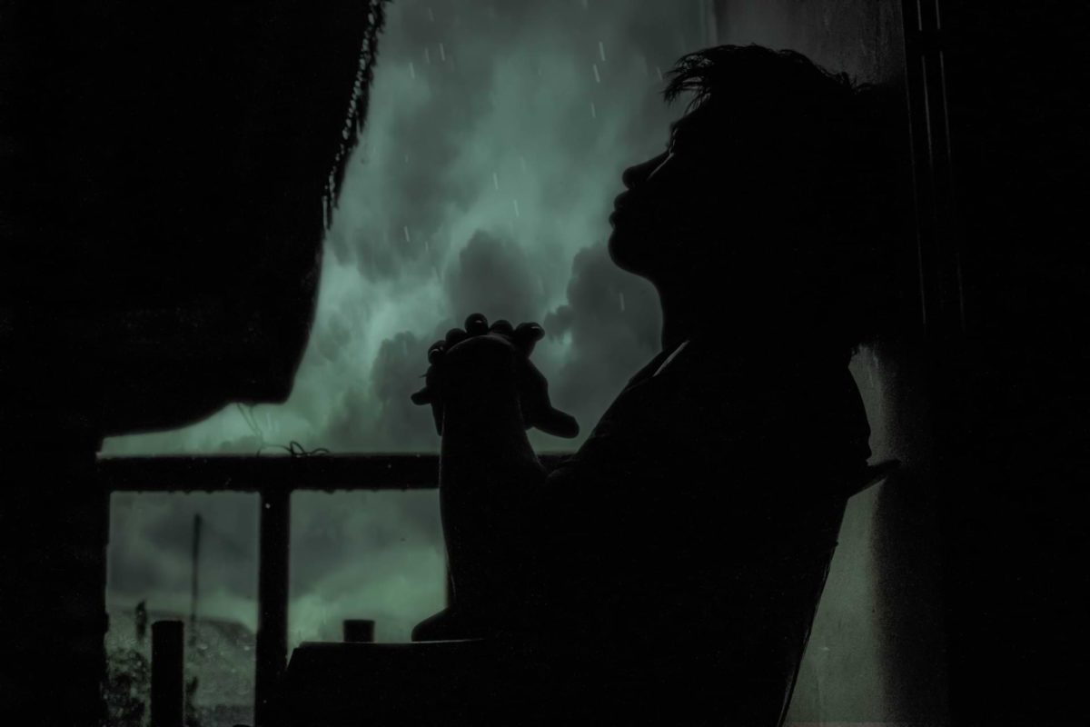 Dark silhouette of a young man sitting alone on a small balcony, looking up at the raining sky—presumably using his solitude to get to know himself better.