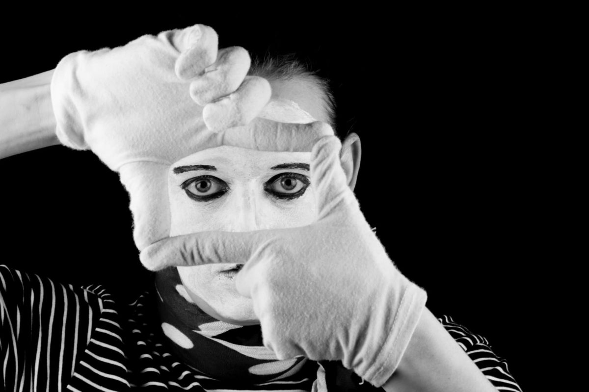 A mime wearing face paint, gloves and a striped shirt, looking through a rectangle made with his fingers from a dark background—figurative of one being lead astray toward societal games.