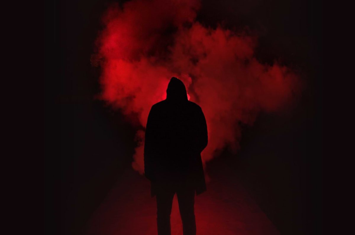 A dark, hooded silhouette facing away from the camera and toward a cloud of smoke, billowing out of a red flare—emblematic of one who feels like the one and only; like the entire world is against them.