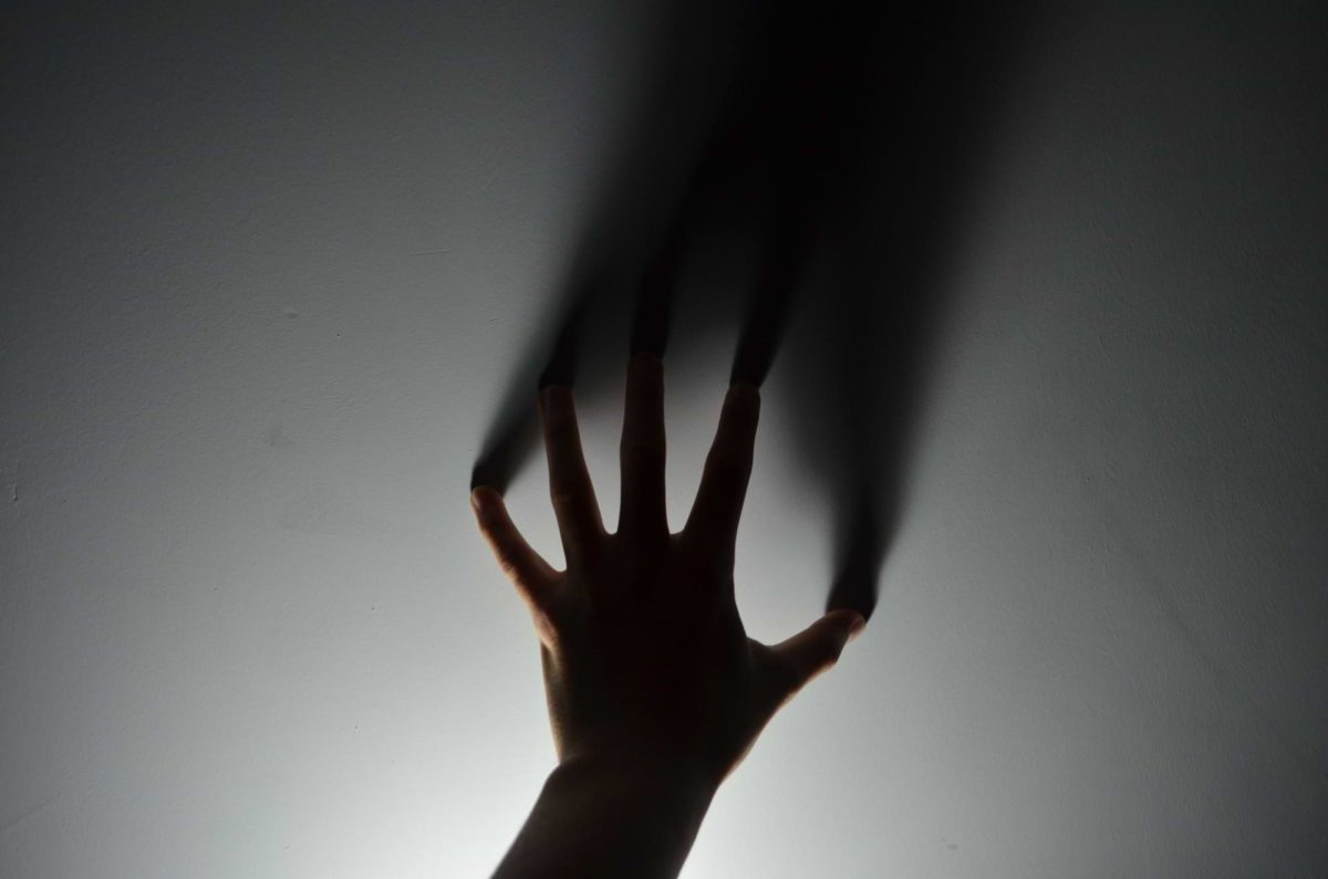 Dark silhouette of a hand spread with its fingertips touching a white wall; its sole light source emanating from below, casting a long shadow above—a depiction of the shadow within us all, and the voice in our heads which speaks from within.